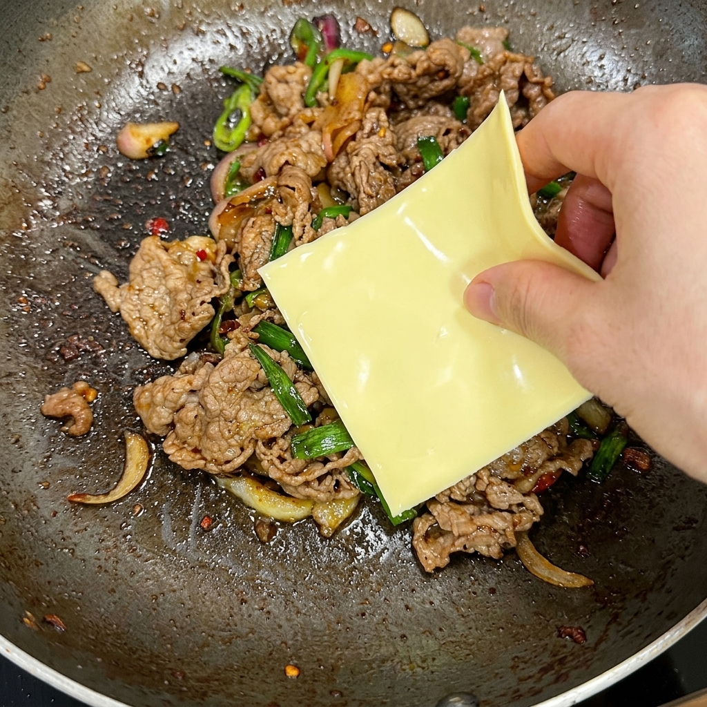 Una mano colocando una loncha cuadrada de queso amarillo sobre la mezcla caliente de carne y chiles en una sartén.