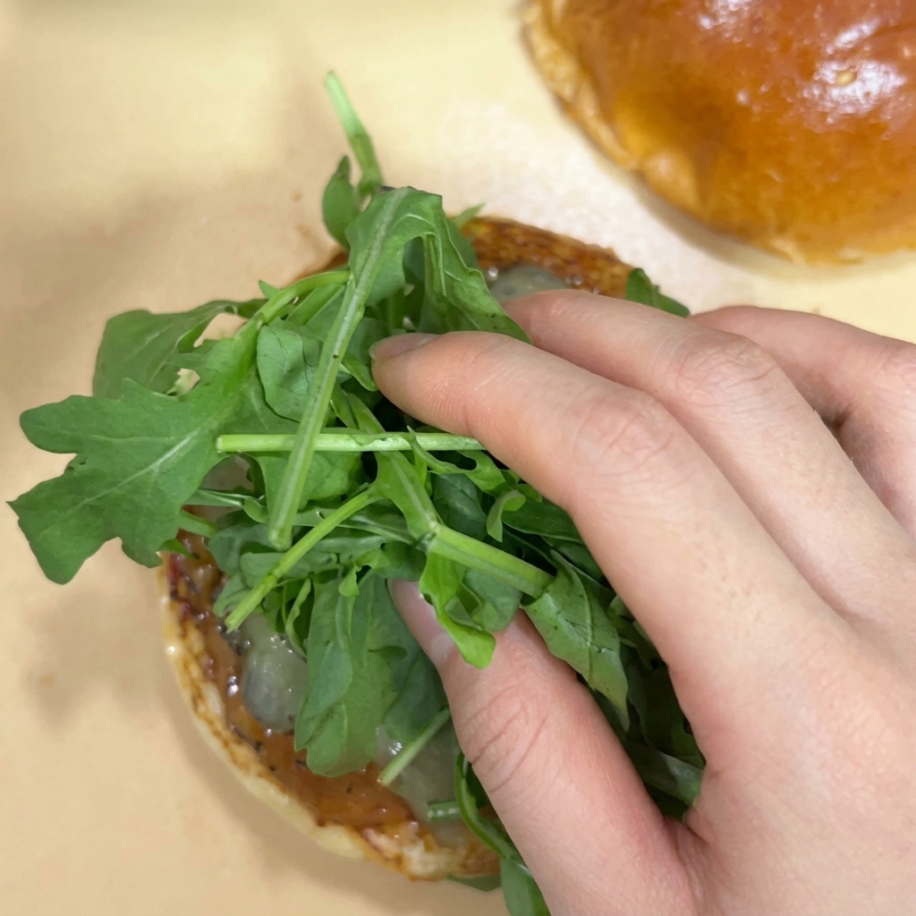 Una mano colocando un manojo fresco de hojas de rúcula verde sobre los pepinillos en la mitad de un pan de hamburguesa tostado.
