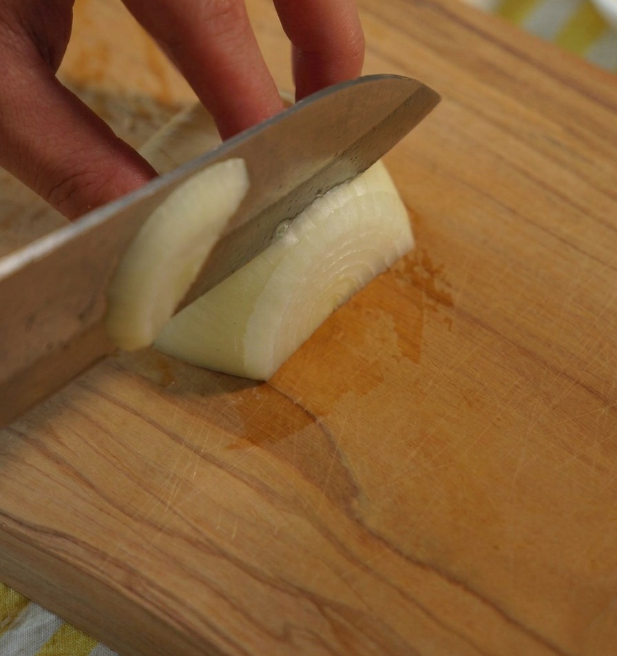 Primer plano de una mano cortando una cebolla blanca en trozos uniformes sobre una tabla de madera con un cuchillo de chef.
