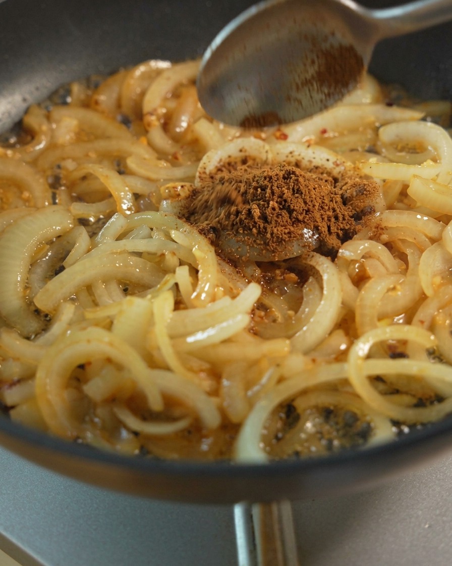 Láminas de cebolla sofriéndose en una sartén mientras se añade azúcar moreno y condimentos líquidos para el caramelizado.