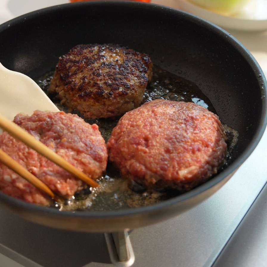Tres hamburguesas gruesas de ternera cocinándose en la sartén, con una volteada mostrando una costra oscura y caramelizada.