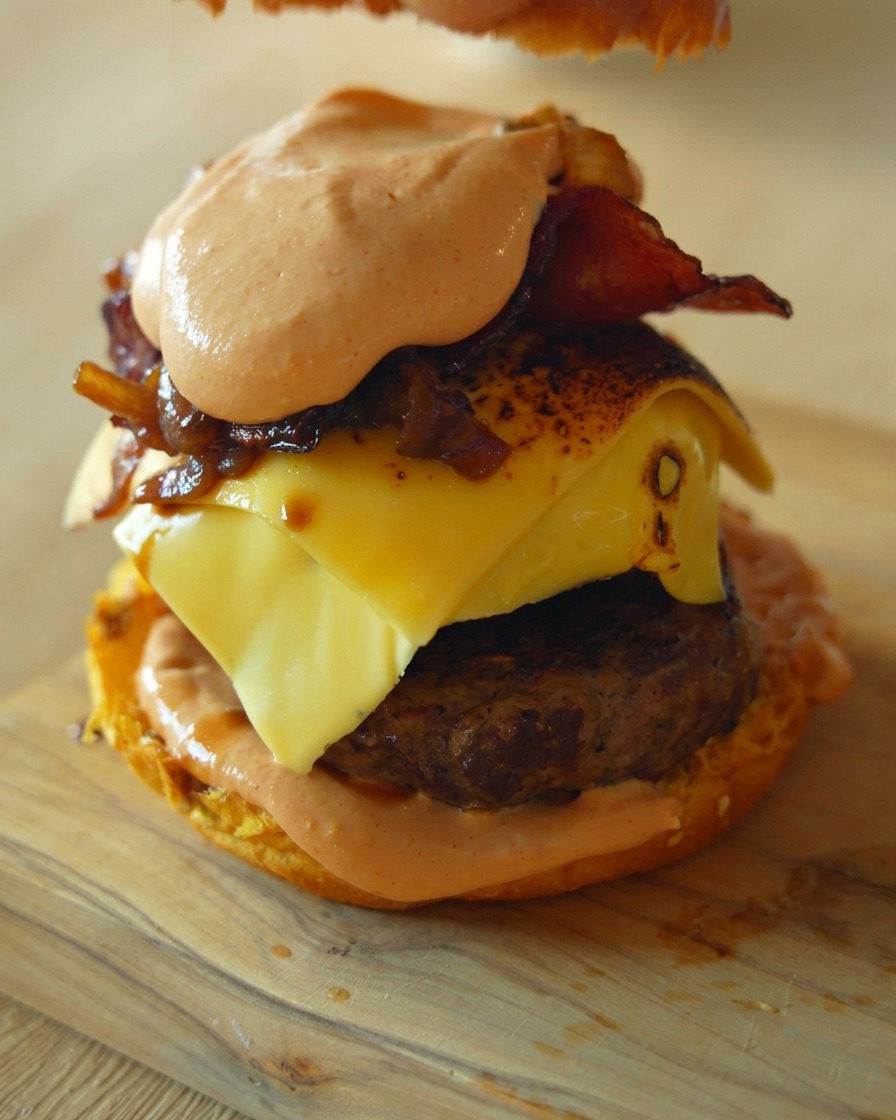 Un primer plano mostrando una mano colocando la tapa del pan sobre una hamburguesa gourmet alta con todos sus ingredientes.