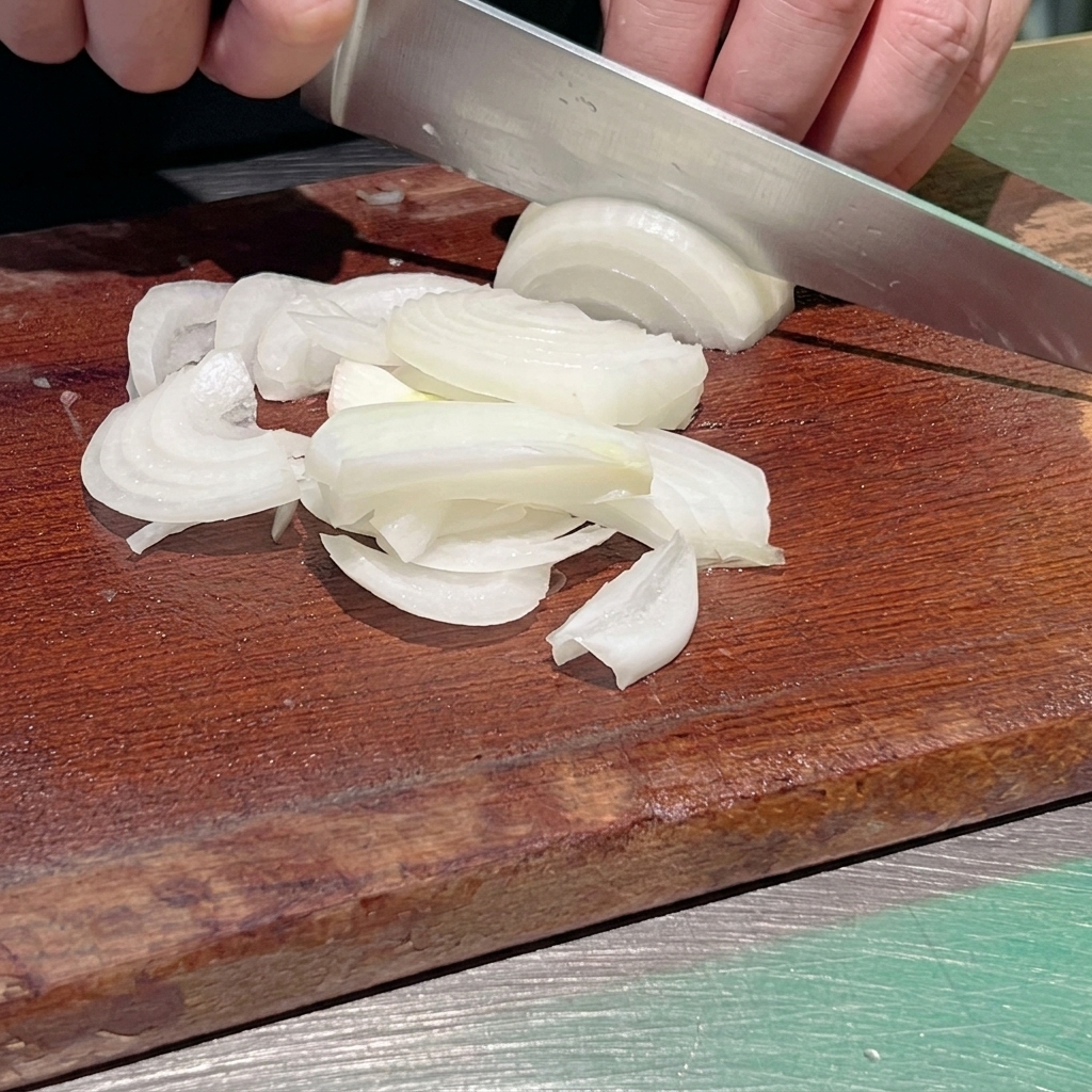Un plano de cerca de un chef usando un cuchillo afilado para cortar una cebolla blanca en tiras finas sobre una tabla de picar de madera oscura.