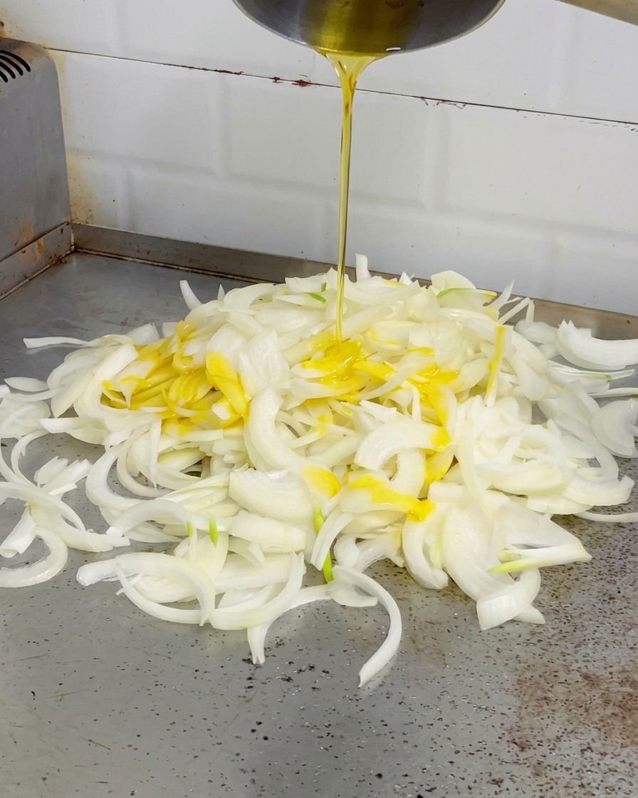 Mantequilla amarilla derretida siendo vertida desde una olla plateada sobre un montón de cebollas blancas en una plancha.