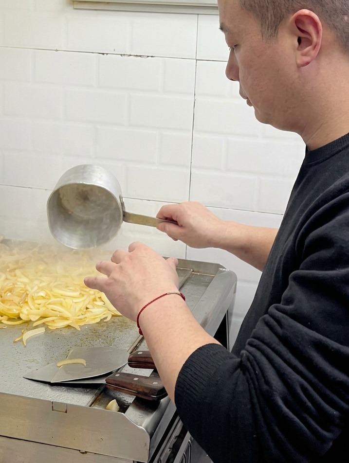 Un chef vertiendo agua desde un pequeño cazo de metal sobre un montón de cebollas que se ablandan en una plancha comercial.