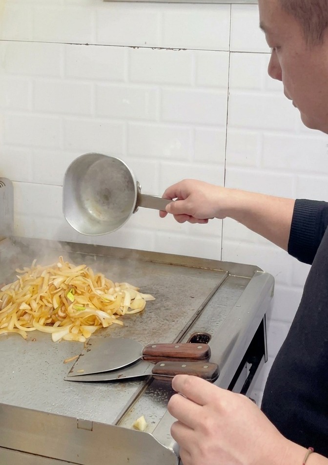 Un chef añadiendo más agua a un montón de cebollas de color marrón claro, parcialmente caramelizadas, en la superficie de una plancha.