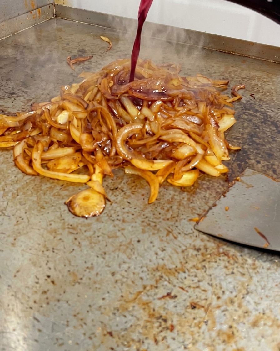 Vino tinto siendo vertido desde una botella sobre un montón de cebollas profundamente doradas en una plancha.