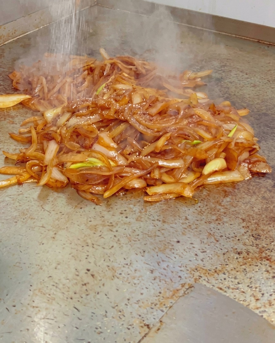 Sal fina siendo espolvoreada sobre un montón de cebollas caramelizadas, brillantes y humeantes, en una plancha caliente.