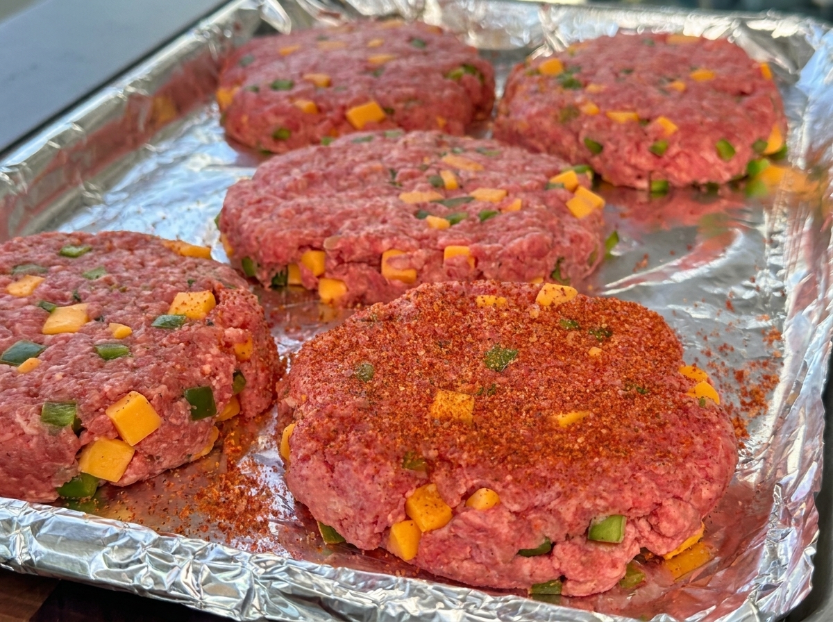 Carnes de hamburguesa crudas formadas con cubos de queso y pimientos visibles, sazonadas con polvo BBQ rojo en una bandeja.