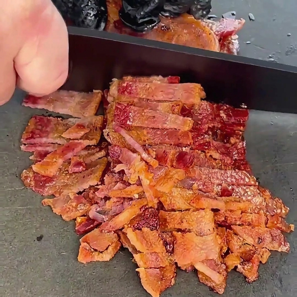 Tiras de tocino cocidas y crujientes siendo picadas finamente con un cuchillo de chef negro sobre una superficie oscura.