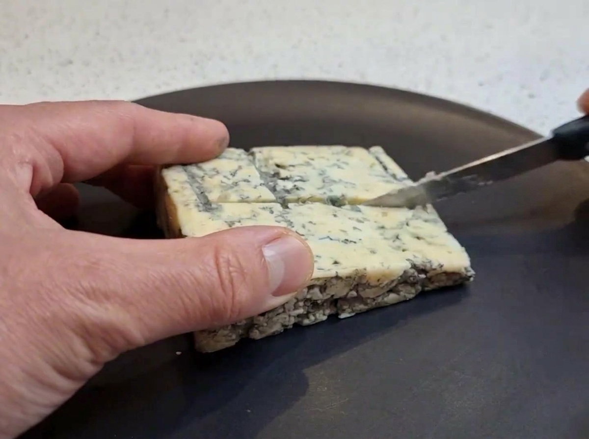 Una mano usando un cuchillo pequeño para cortar un bloque cuadrado de queso azul en trozos más pequeños sobre un plato oscuro.