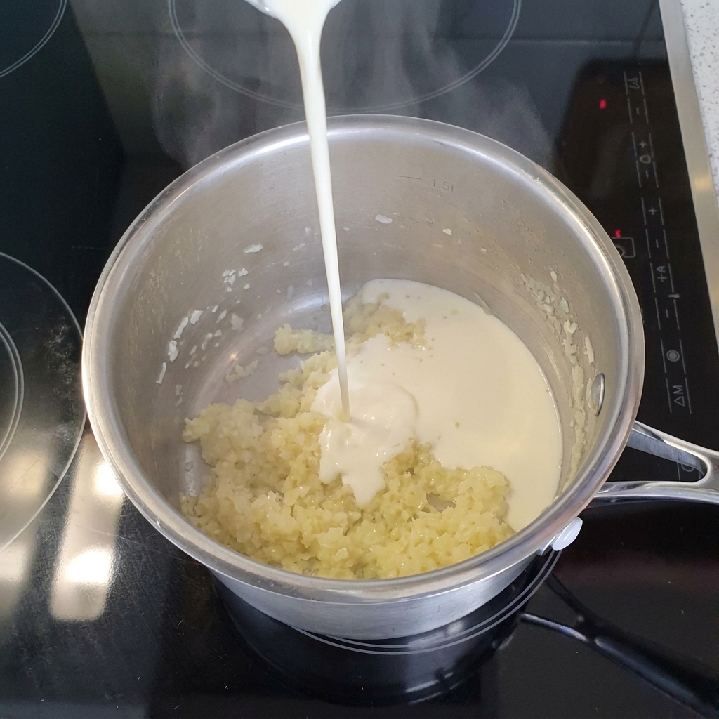 Un chorro de crema ligera líquida vertiéndose en una olla de acero inoxidable llena de cebolla y ajo picados y cocidos.