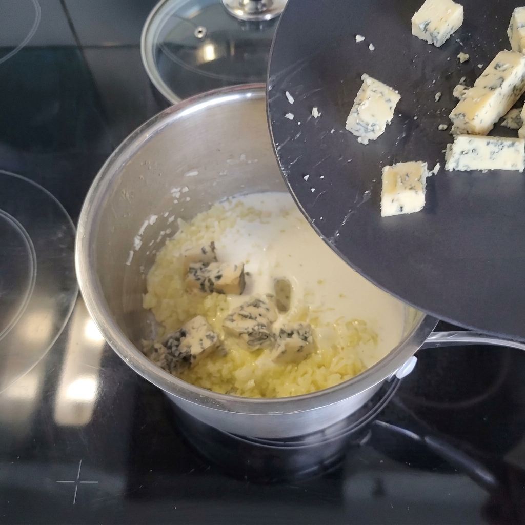 Trozos de queso azul deslizándose desde un plato negro inclinado hacia una cacerola que contiene crema y aromáticos salteados.