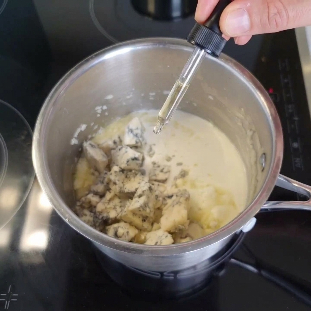Una mano usando un gotero de vidrio para añadir salsa inglesa oscura en una olla de metal llena de crema ligera y trozos de queso azul.