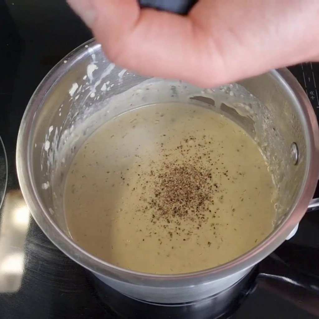 Una mano moliendo pimienta negra fresca con un molinillo directamente sobre una salsa cremosa dentro de una olla de acero inoxidable en una estufa negra.