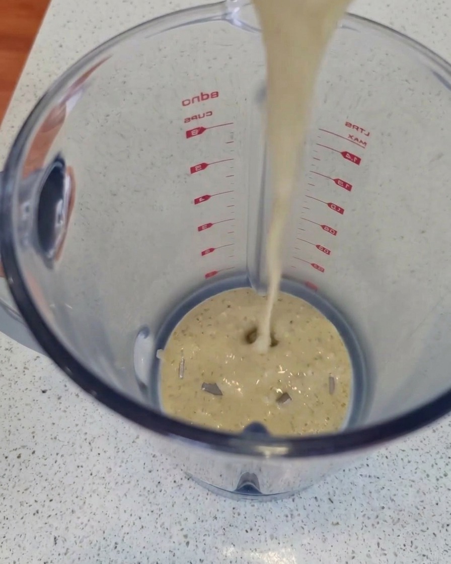 Un flujo de salsa de queso azul cremosa y con motitas vertiéndose desde arriba en la jarra de plástico transparente de una licuadora sobre la encimera de la cocina.