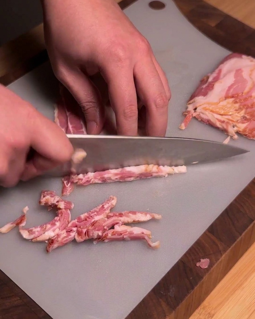 Un chef utilizando un cuchillo afilado para cortar tiras de tocino crudo en trozos uniformes sobre una tabla de cortar gris.