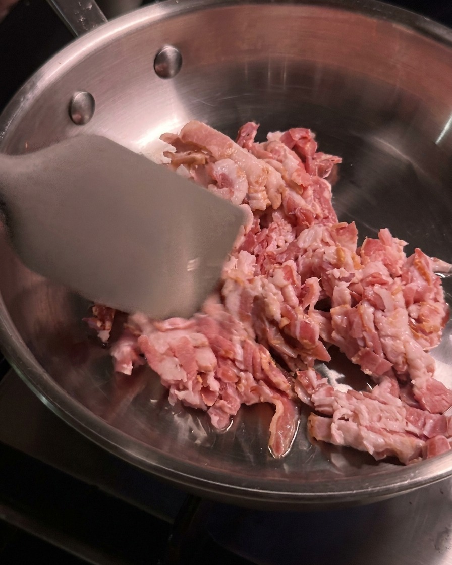 Trocitos de tocino crudo cocinándose y removiéndose con una espátula de metal en una sartén de acero inoxidable caliente.