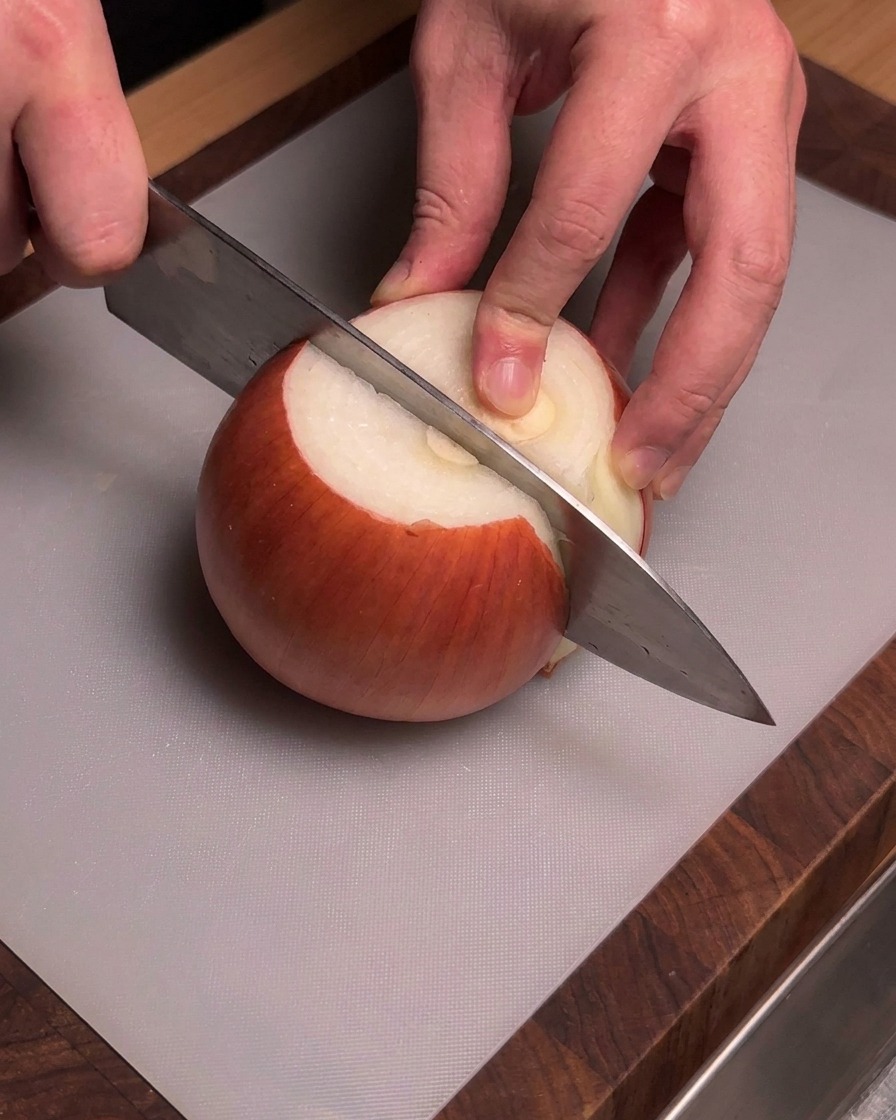 Manos sosteniendo un cuchillo de chef para cortar una cebolla amarilla pelada sobre una tabla de cortar gris.