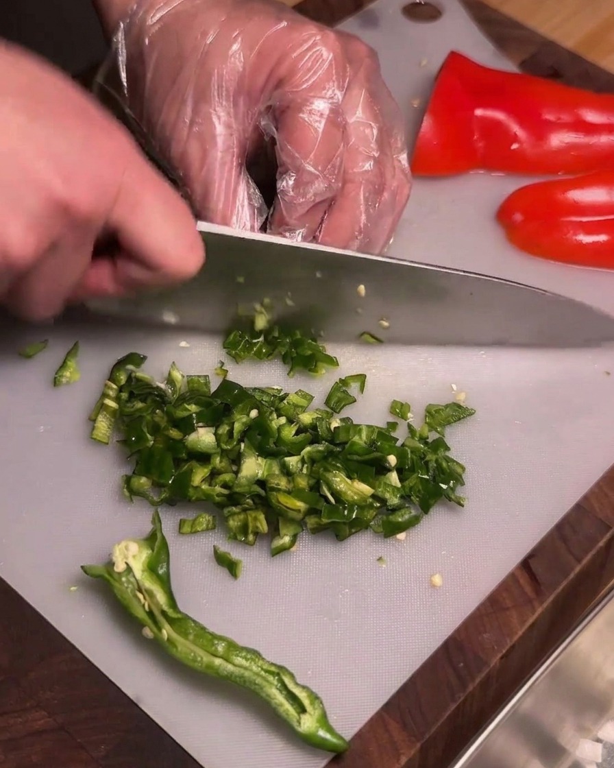 Una persona con guantes de plástico transparente picando finamente chiles verdes sin semillas en una tabla de cortar.