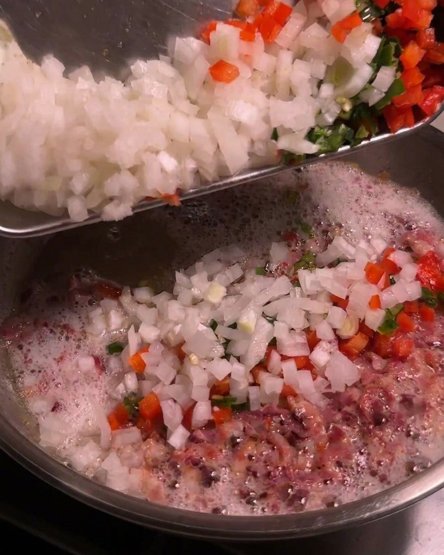 Una tabla con cebolla blanca picada y chiles de colores volcándose en una sartén burbujeante con grasa de tocino.