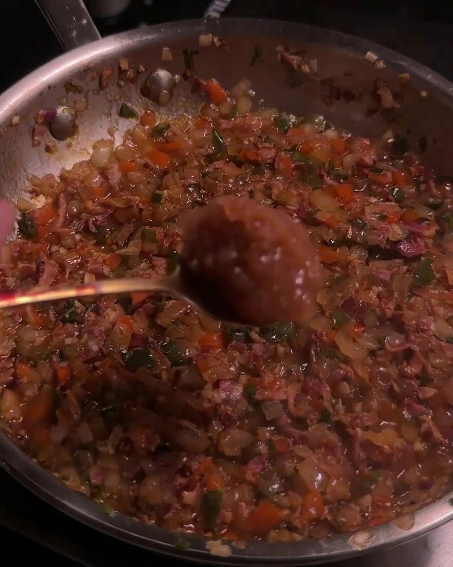 Una cuchara con puré de manzana añadiéndose a una mezcla caramelizada de tocino y chiles en una sartén.