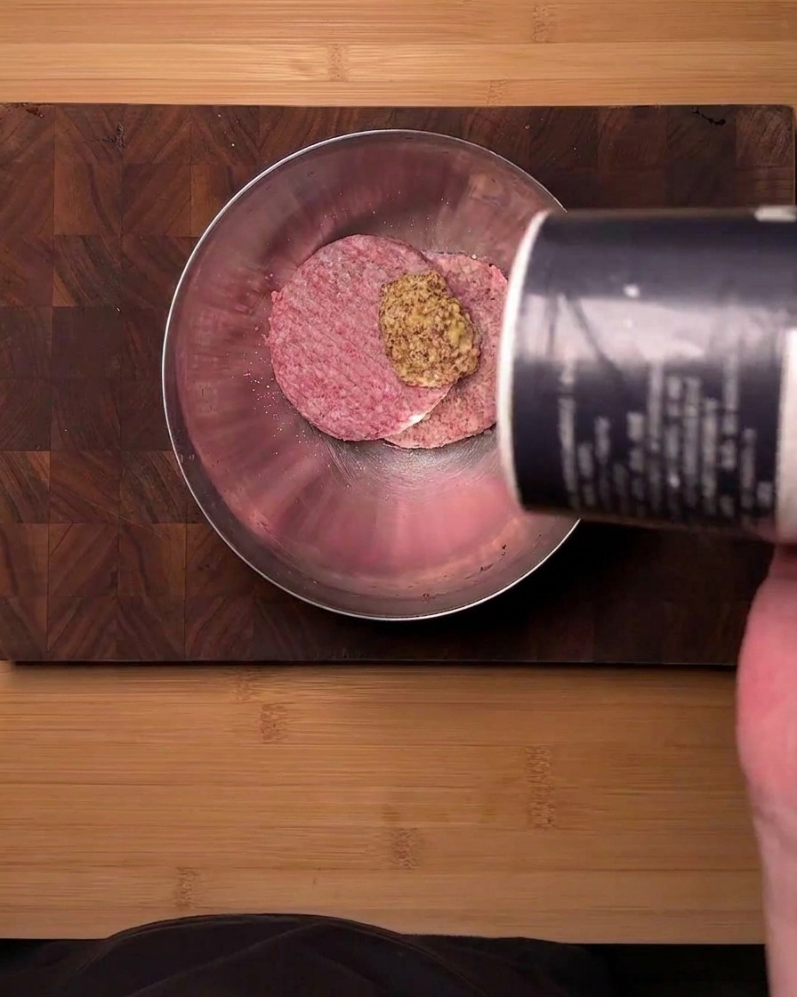 Vista superior de carne picada cruda en un bol de acero inoxidable sazonándose con mostaza en polvo y sal de un recipiente.