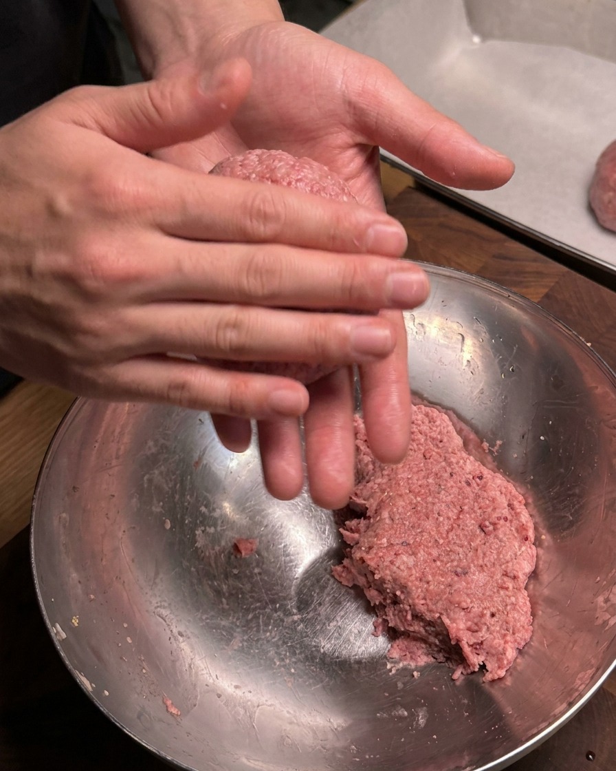 Un primer plano de manos moldeando firmemente una porción de carne picada sazonada cruda en una albóndiga redonda sobre un bol de metal.