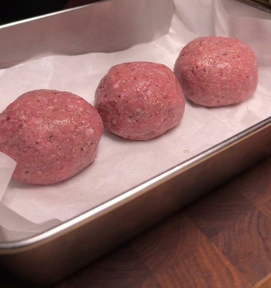 Tres albóndigas de res crudas ordenadas sobre papel pergamino blanco en una bandeja de metal, sus superficies brillando uniformemente con una capa de humo líquido.