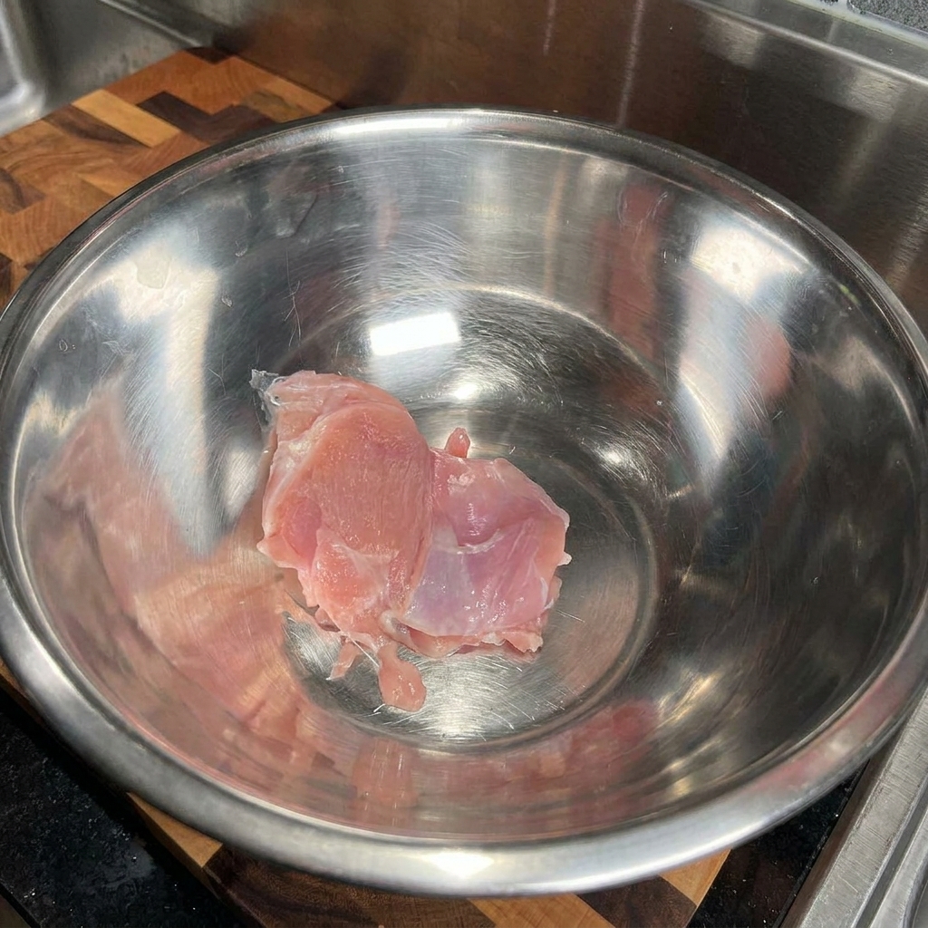 Contramuslo de pollo crudo colocado dentro de un tazón de mezcla grande y vacío de acero inoxidable.
