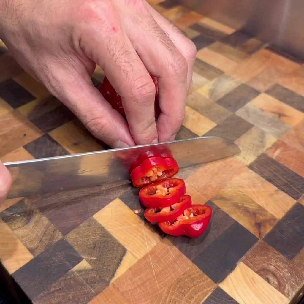 Una persona usando un cuchillo de cocina para cortar chiles rojos frescos en rodajas sobre una tabla de cortar de madera.