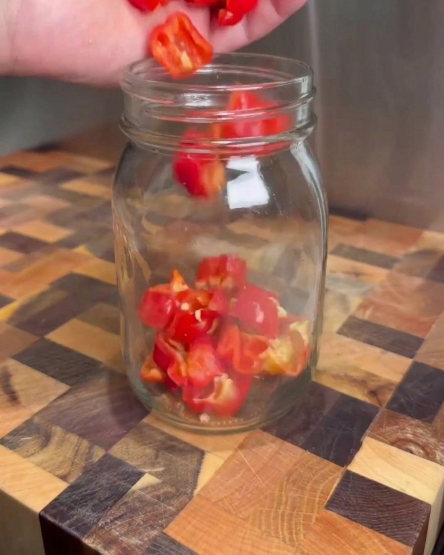 Chiles rojos picados cayendo dentro de un frasco de vidrio transparente sobre una tabla de cortar de madera.