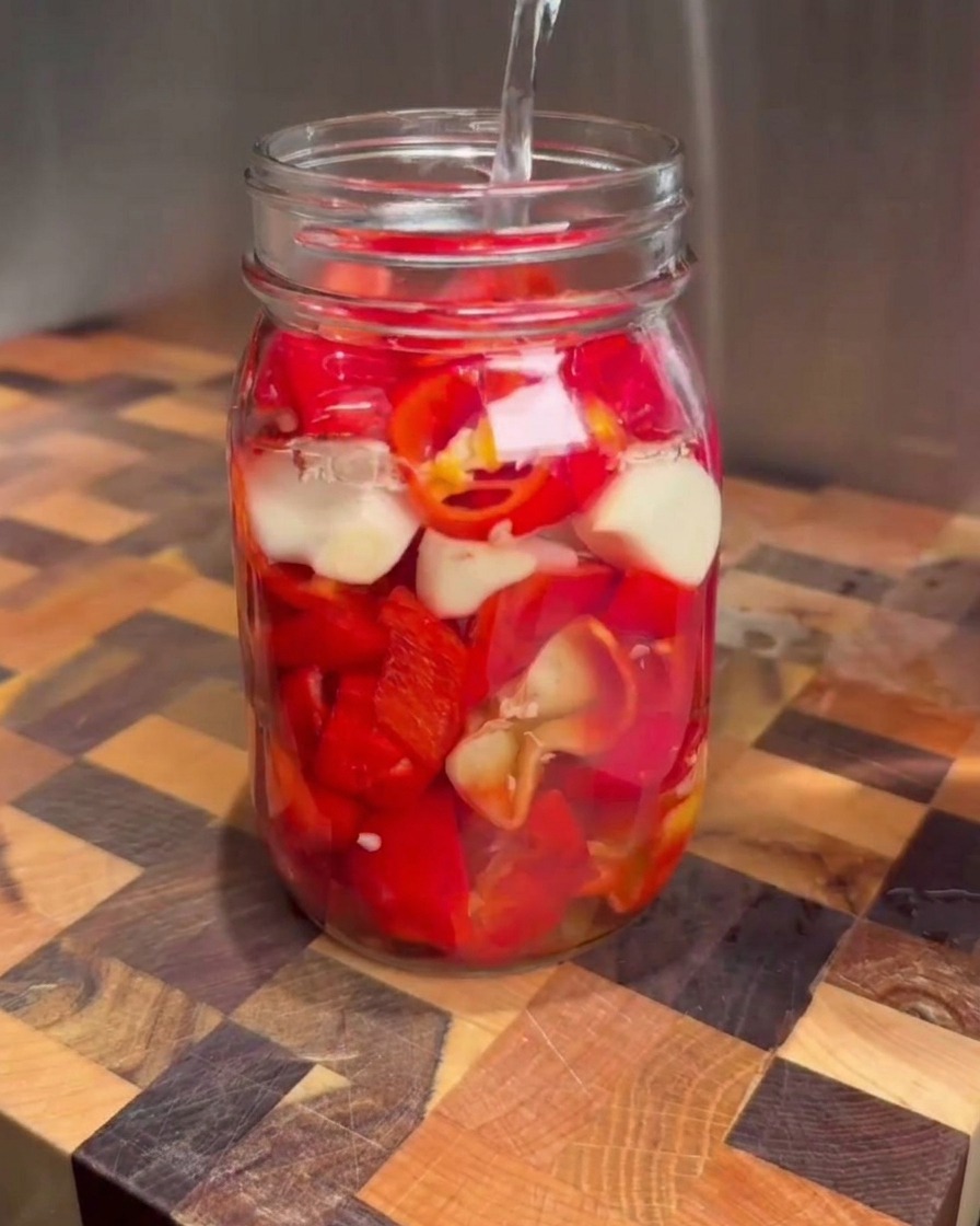 Salmuera clara vertiéndose desde una taza medidora en un frasco de vidrio lleno de chiles rojos picados y dientes de ajo.
