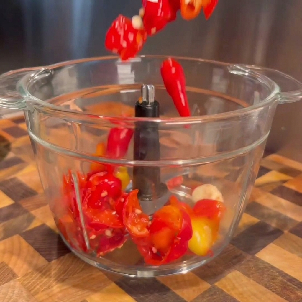 Chiles fermentados rojos y dientes de ajo escurridos cayendo en el tazón transparente de un procesador de alimentos.