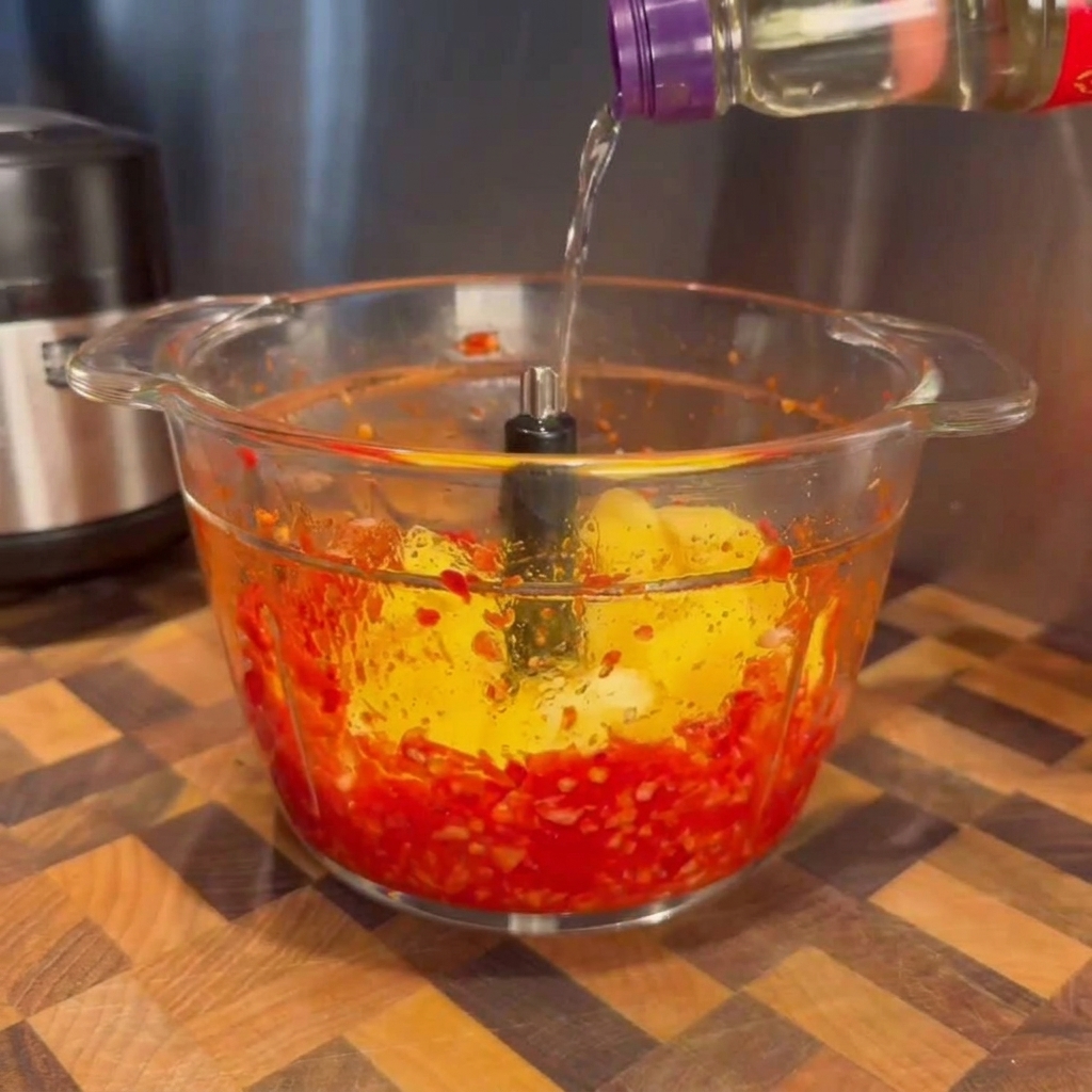 Líquido transparente vertiéndose desde una botella en el tazón de vidrio de un procesador de alimentos que contiene chiles rojos licuados y trozos de mango amarillo brillante.
