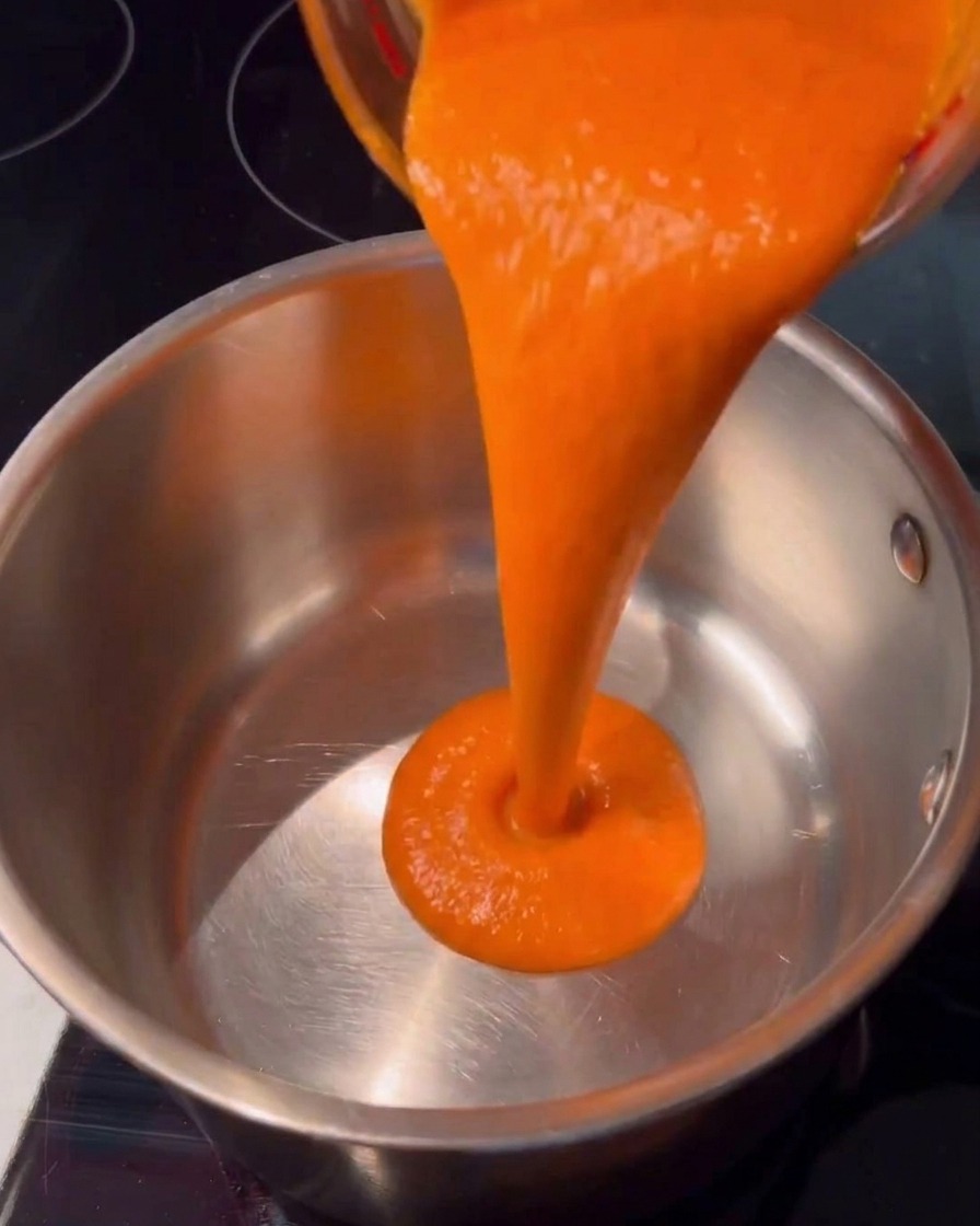 Salsa picante de color naranja brillante vertiéndose perfectamente desde una taza de vidrio en una cacerola limpia de acero inoxidable sobre una estufa oscura.