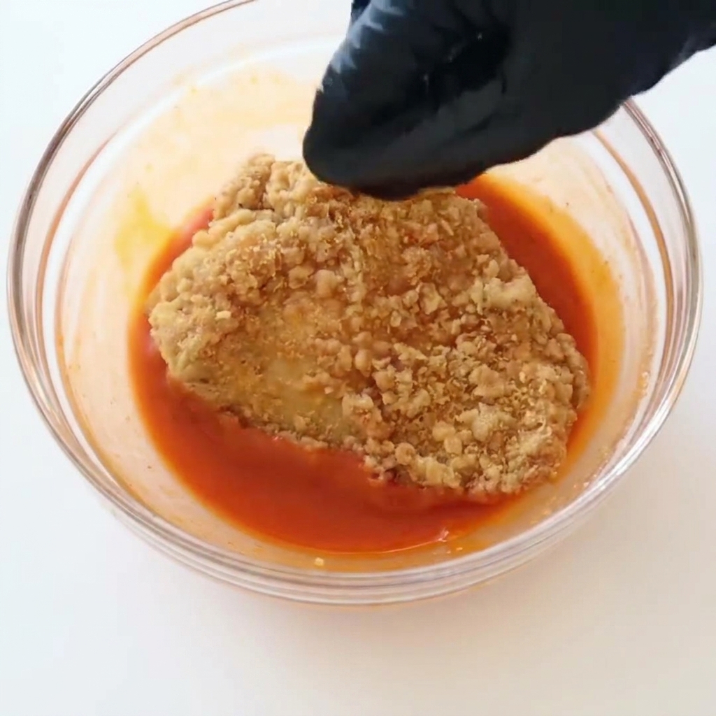 Una mano con guante negro sumergiendo un filete de pollo frito crujiente en un bol de salsa búfalo roja.