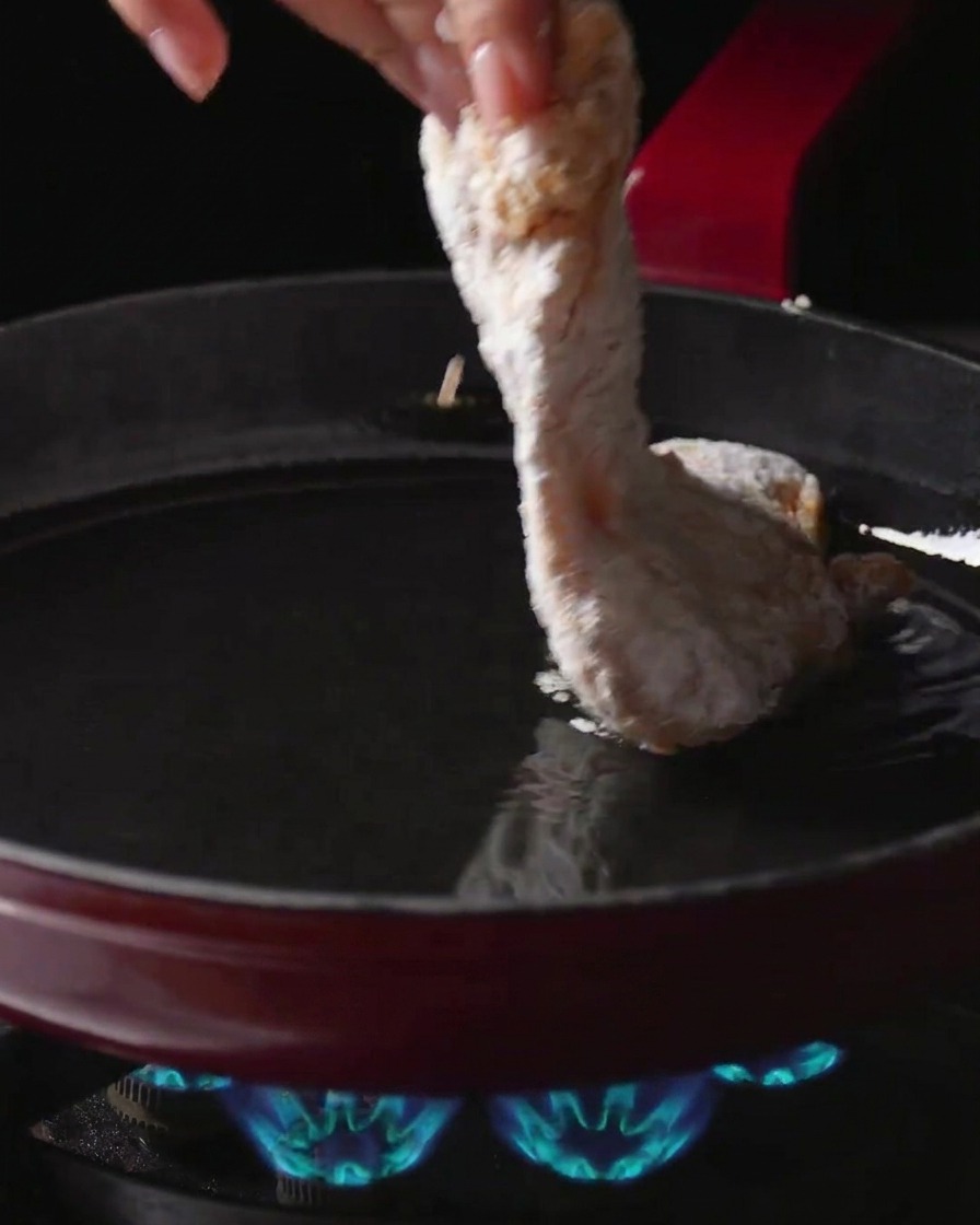 Una mano colocando un contramuslo de pollo enharinado en una sartén con aceite caliente sobre una hornilla de gas encendida.