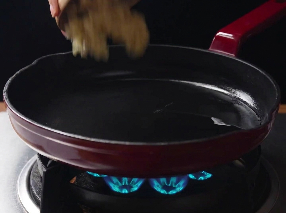 Ajo picado vertiéndose desde un tazón pequeño en una sartén caliente con el aceite restante sobre una estufa de gas encendida.