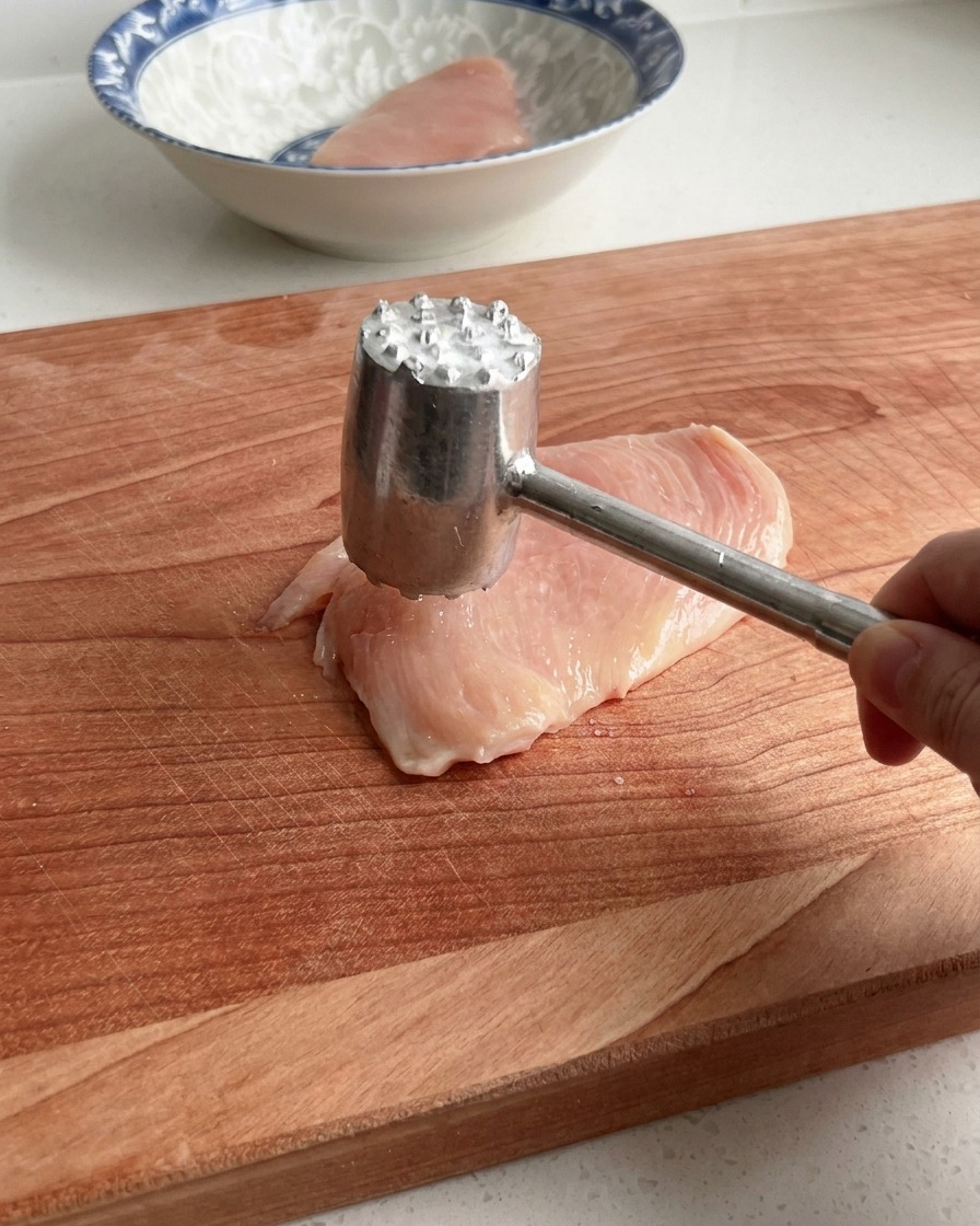 Una mano usando un mazo ablandador de carne para golpear suavemente una pechuga de pollo cruda sobre una tabla de cortar de madera.