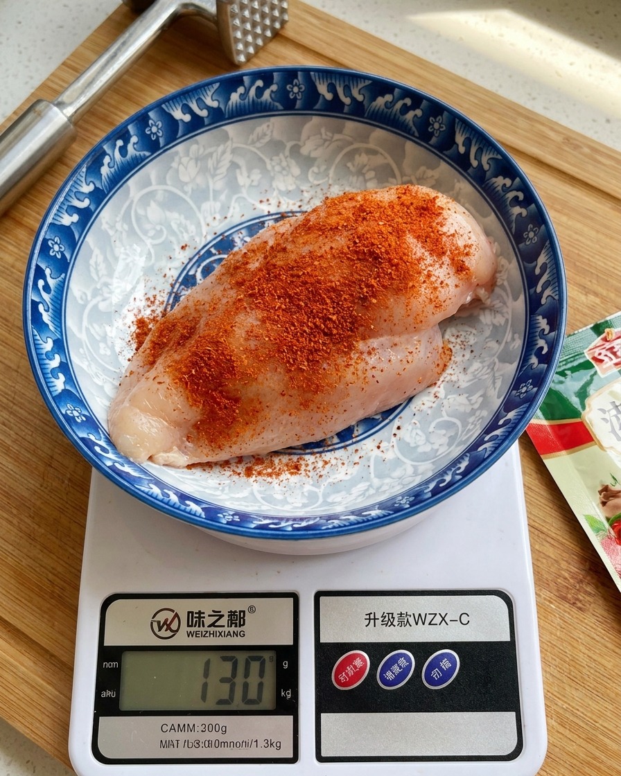 Una pechuga de pollo cruda en un tazón de cerámica decorado sobre una báscula digital de cocina, espolvoreada con sazonador de color naranja rojizo.