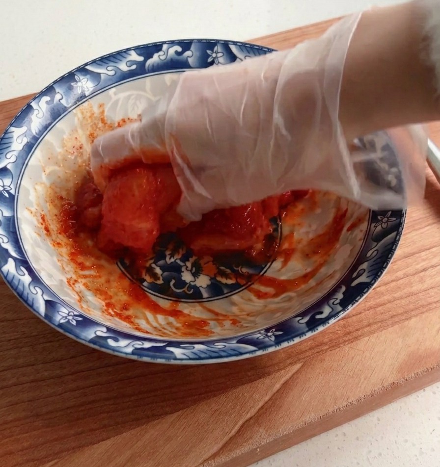 Una mano con guante cubriendo y mezclando bien una pechuga de pollo cruda con un marinado espeso y rojo dentro de un tazón decorado.