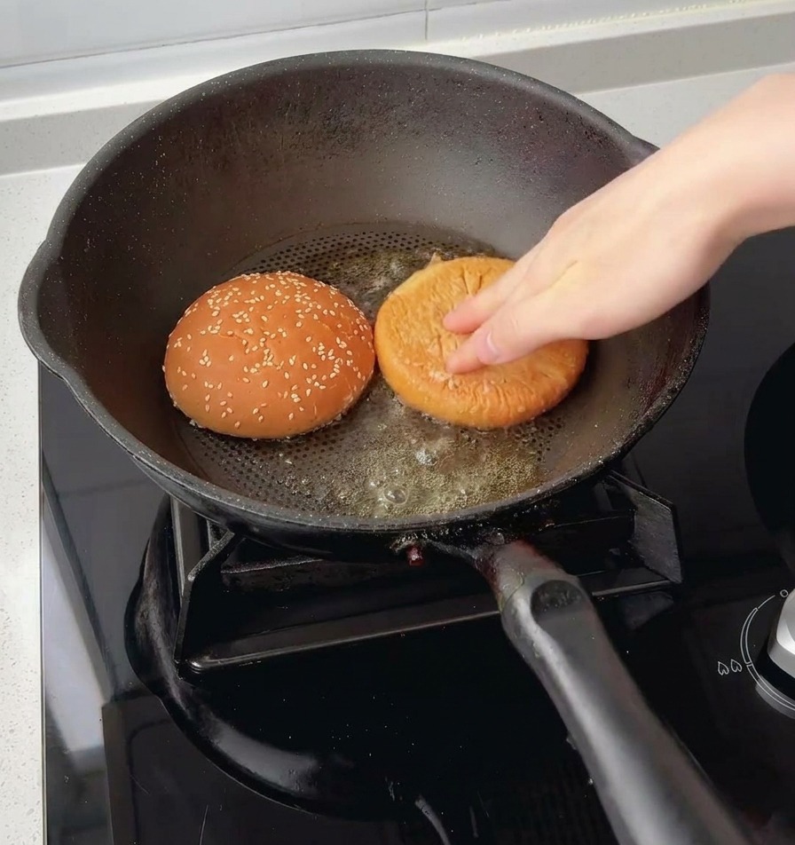 Una mano presionando las dos mitades de un pan de hamburguesa con ajonjolí boca abajo sobre la mantequilla derretida en una sartén.