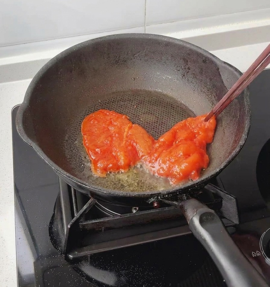 Unas pinzas o palillos chinos colocando una pechuga de pollo roja y completamente marinada en una sartén negra ligeramente engrasada.
