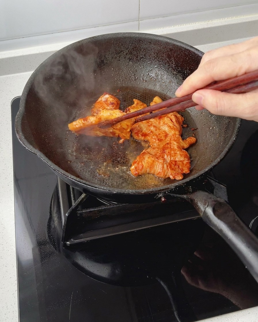 Dorando trozos de pechuga de pollo marinada en una sartén mientras se les da la vuelta con palillos, mostrando una cobertura glaseada y caramelizada.