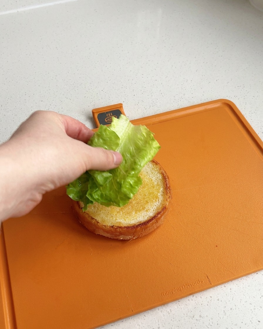 Una mano colocando una hoja de lechuga verde fresca y limpia sobre la base dorada de un pan de hamburguesa de ajonjolí en una tabla de cortar naranja.