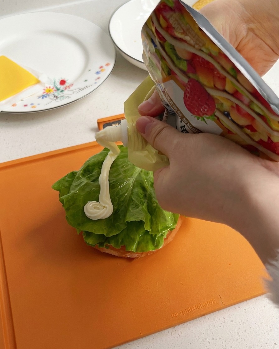 Mayonesa blanca y cremosa siendo exprimida en una línea gruesa sobre una hoja de lechuga verde en la base de un pan de hamburguesa.