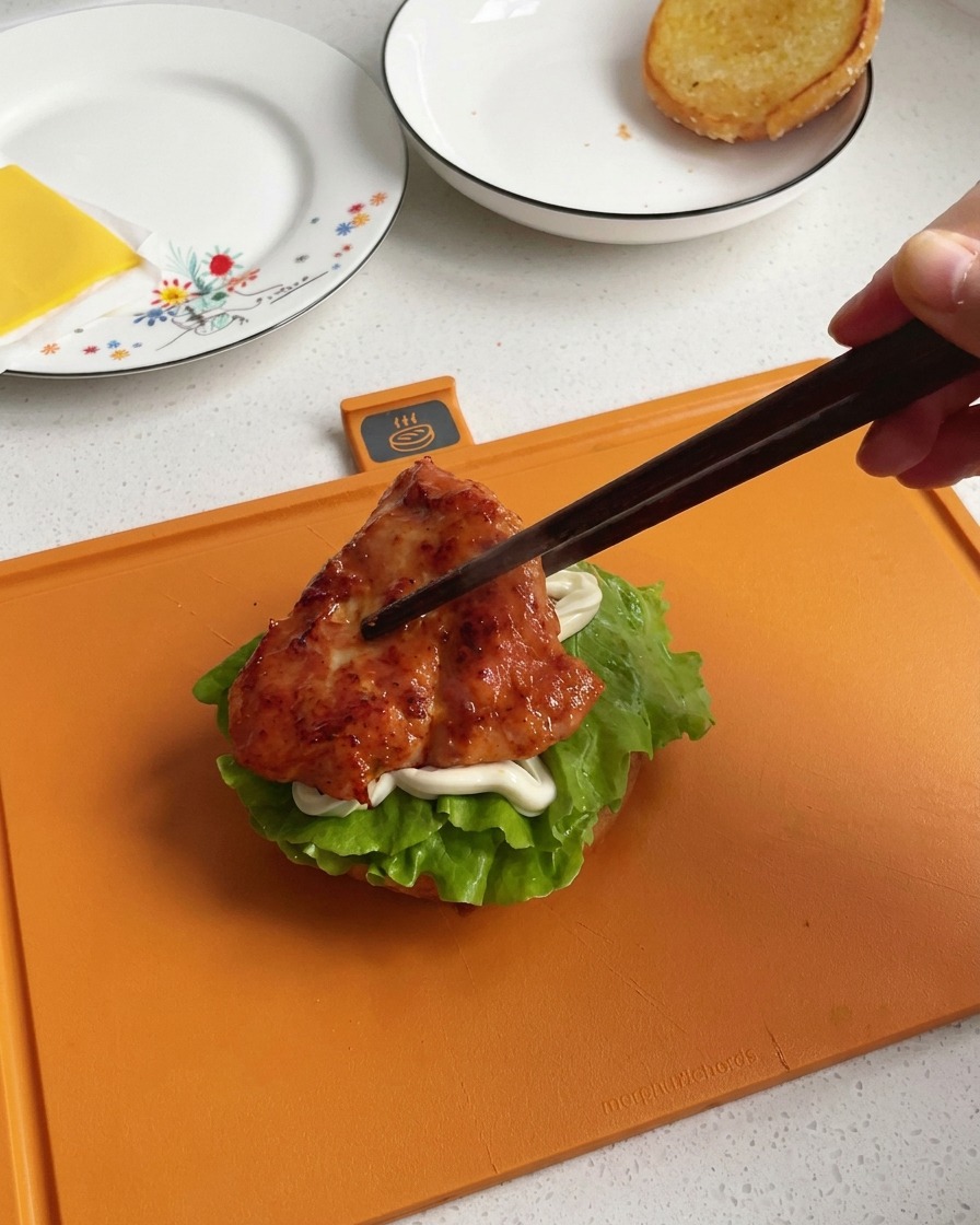 Una mano usando palillos oscuros de madera para pasar un trozo de pollo a la sartén glaseado y muy caramelizado al armado de la hamburguesa.