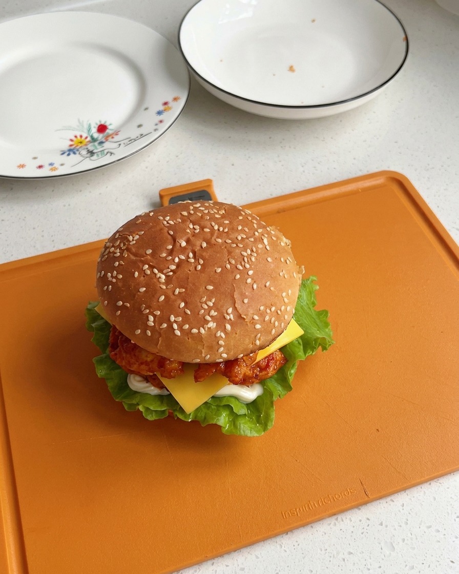Una hamburguesa de pollo Orleans completamente armada y coronada con un pan con ajonjolí, mostrando capas visibles de lechuga, pollo y una rebanada de queso derritiéndose.