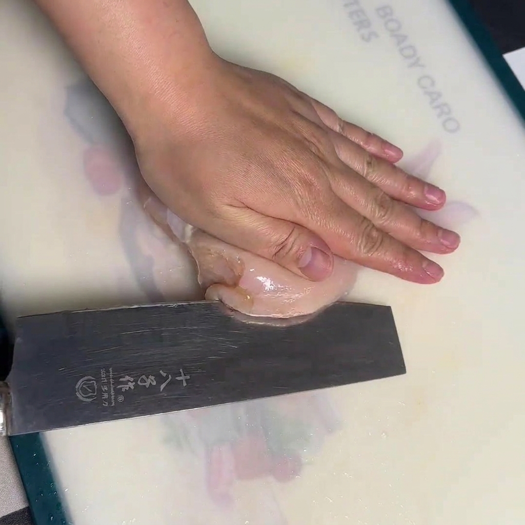 Una mano sosteniendo una pechuga de pollo cruda sobre una tabla de cortar blanca mientras la filetea horizontalmente con un cuchillo de chef.